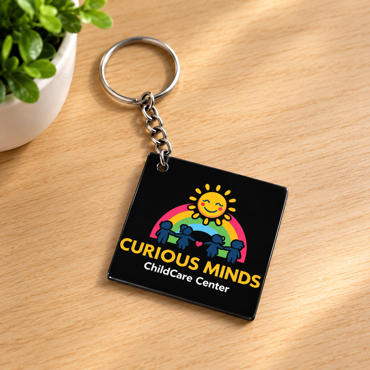 A square black acrylic keychain with a colorful graphic of a smiling sun, rainbow, and child silhouettes, with the text 'CURIOUS MINDS ChildCare Center' in yellow and white, resting on a light wood surface next to a small potted plant.