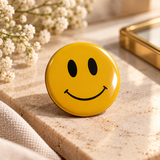 Glossy yellow smiley pin badge displayed on a cream marble surface with elegant styling