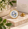 A round enamel pin with a gold border and white face displaying the CA INDIA logo, shown on a stone slab next to its gold clasp and a branded box in a styled setting.