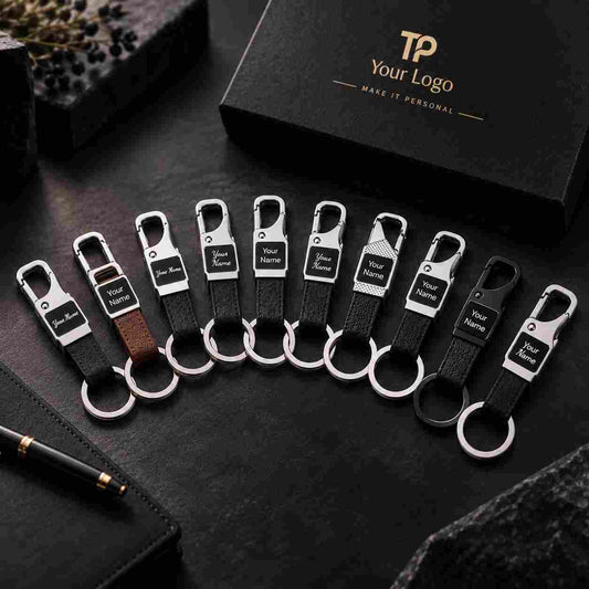 A set of personalized keychains arranged in a fan shape on a dark textured surface, with a branded black gift box in the background. The keychains feature silver-tone metal clasp ends, black or brown leather straps, and a small rectangular black tag with placeholder text that reads 'Your Logo'.