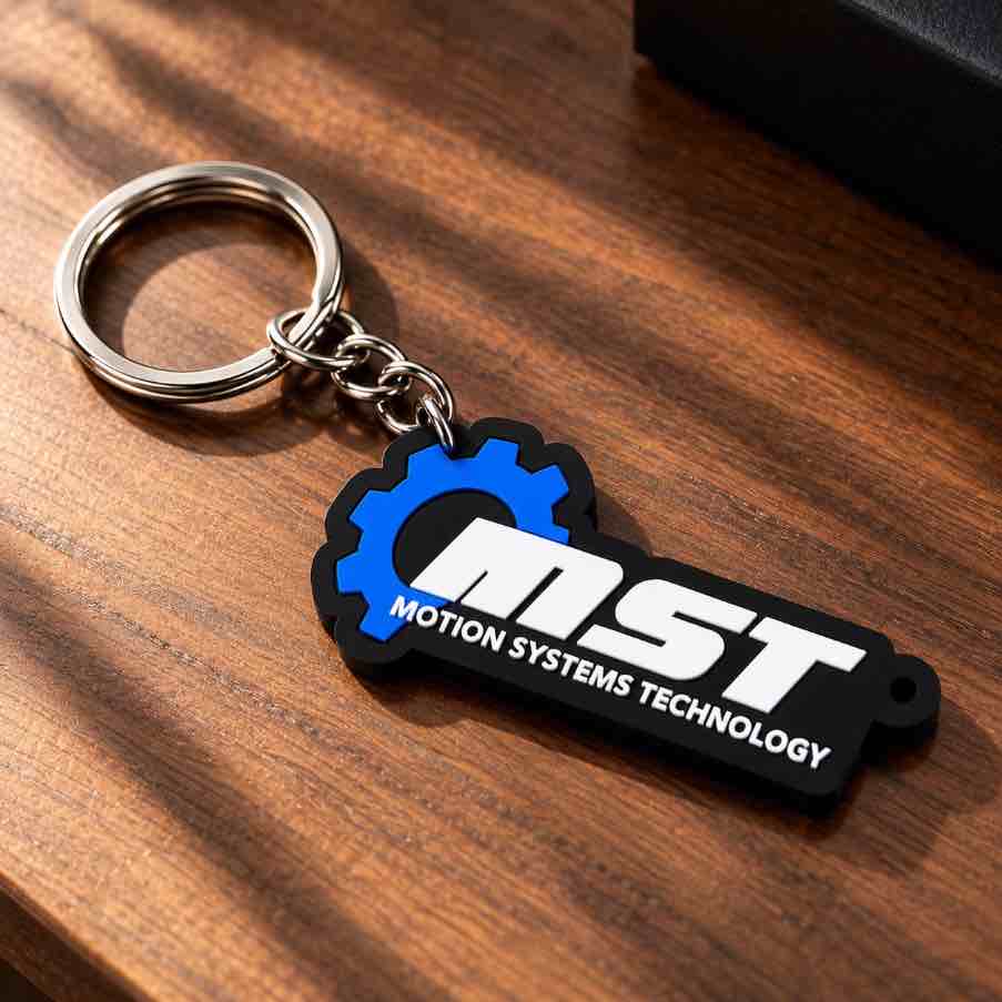 A black and blue rubber keychain with a white 'MST MOTION SYSTEMS TECHNOLOGY' logo, attached to a silver metal split ring, resting on a wooden surface.