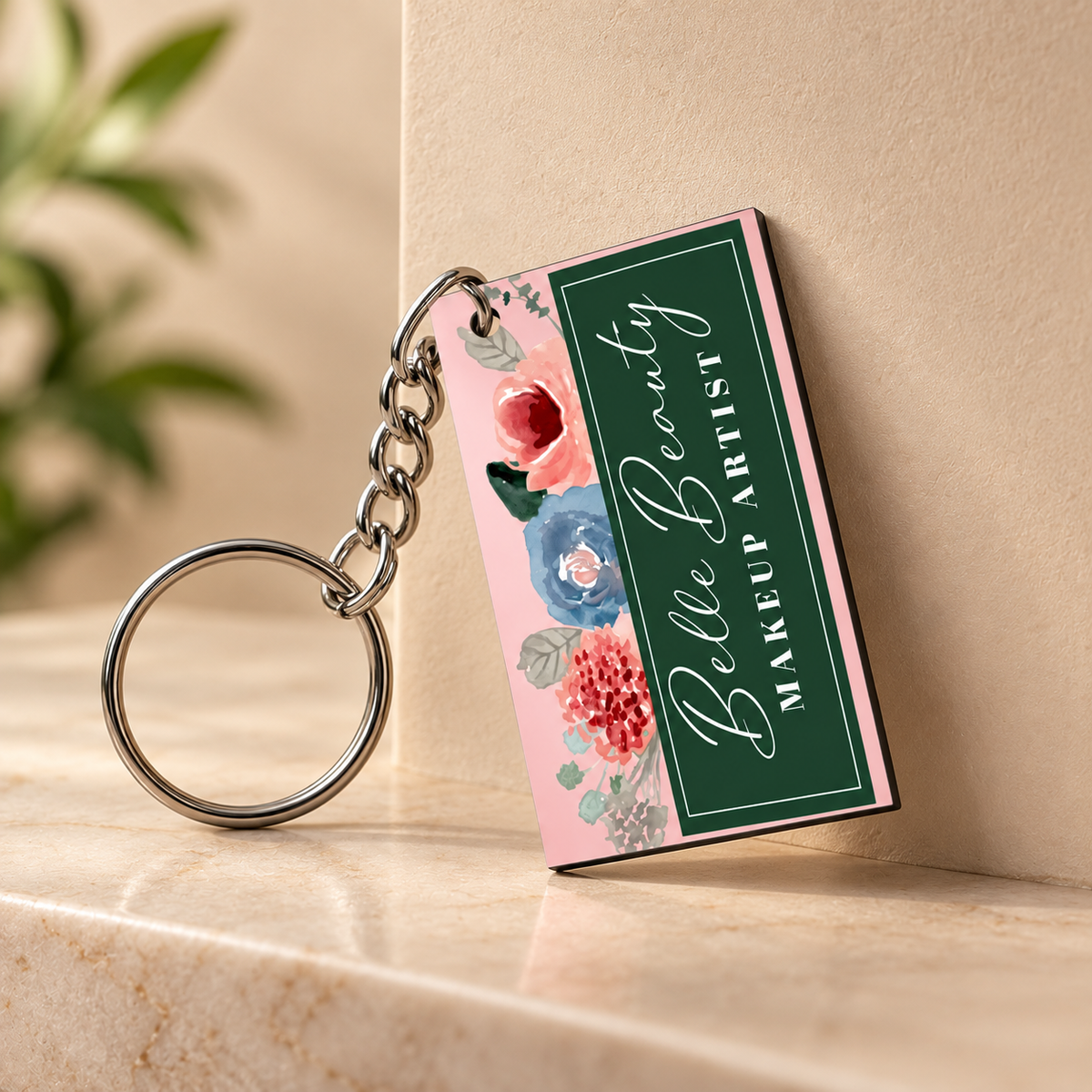 A rectangular acrylic keychain with a silver metal key ring and chain, leaning against a beige wall on a marble surface. The keychain features a pink background with watercolor flowers and a dark green vertical panel with white script reading 'Belle Beauty MAKEUP ARTIST'.