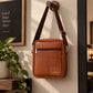 A brown leather crossbody bag hanging from a wooden peg on a beige wall, with a potted plant and framed sign in the background.