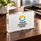 A clear acrylic name block sign with a white insert displaying a colorful circular logo and the placeholder text 'YOUR DESIGN HERE', placed on a wooden desk with a blurred plant and notebook in the background.