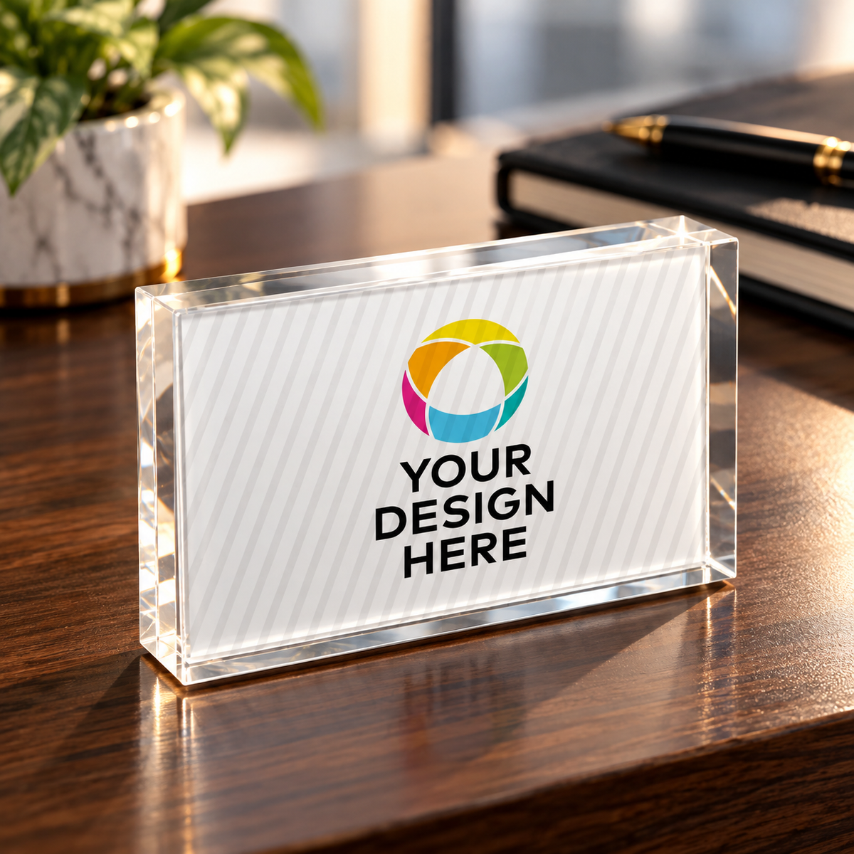 A clear acrylic name block sign with a white insert displaying a colorful circular logo and the placeholder text 'YOUR DESIGN HERE', placed on a wooden desk with a blurred plant and notebook in the background.