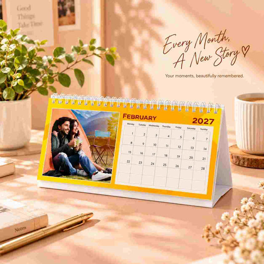 A personalized spiral-bound monthly desk calendar stands on a tabletop. The calendar page shows February 2027 with a printed photo of a couple sitting by an orange tent.
