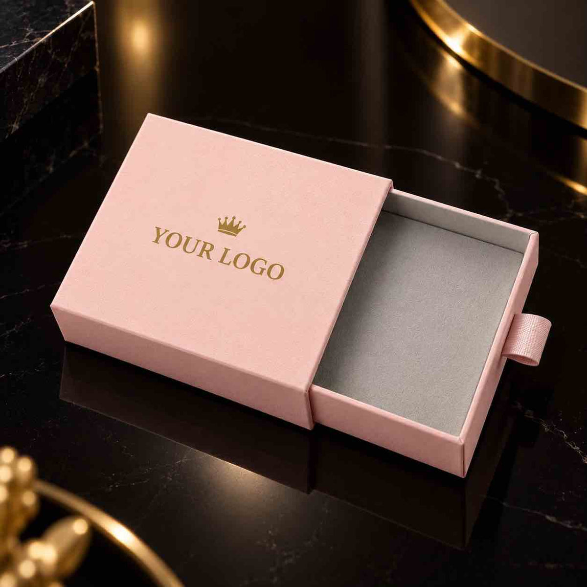 A pink rectangular jewelry box with the lid partially open, showing a gray velvet interior. The lid is printed with a small gold crown icon and the words 'YOUR LOGO' in gold.