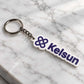 White Kelsun acrylic keychain with purple logo and lettering placed diagonally on white marble surface. 