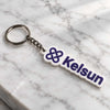 White Kelsun acrylic keychain with purple logo and lettering placed diagonally on white marble surface. 