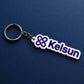 White acrylic Kelsun custom keychain with purple branding displayed diagonally on a deep navy blue surface