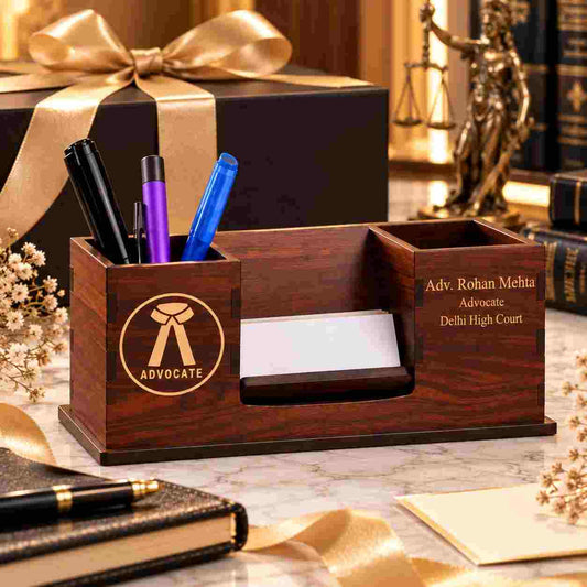 A dark wood desktop organizer with three compartments, engraved with an advocate ribbon logo and the text 'Adv. Rohan Mehta Advocate Delhi High Court', holding pens, a business card slot, and a small tray, styled on a marble surface with a gift box and books in the background.