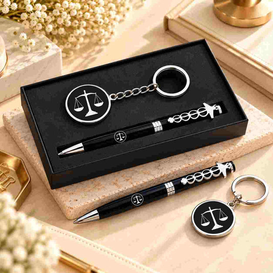 A black and silver pen and keychain set displayed in a black gift box, with additional matching items placed beside it on a beige surface. The set features a pen and keychain with a scale of justice emblem, decorative chain details, and polished silver accents.