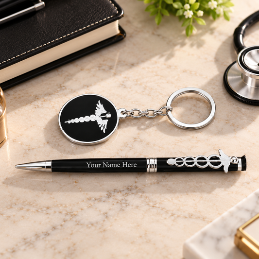 A black and silver medical-themed keychain and pen set on a beige marble desk surface. The round keychain has a black face with a white caduceus symbol, and the pen is black with silver accents, a small caduceus charm, and placeholder text 'Your Name Here'.