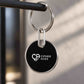 A round black keychain with a silver metal rim and ring, featuring the white Cipher Aces logo, hanging from a dark metal bar against a softly blurred background.