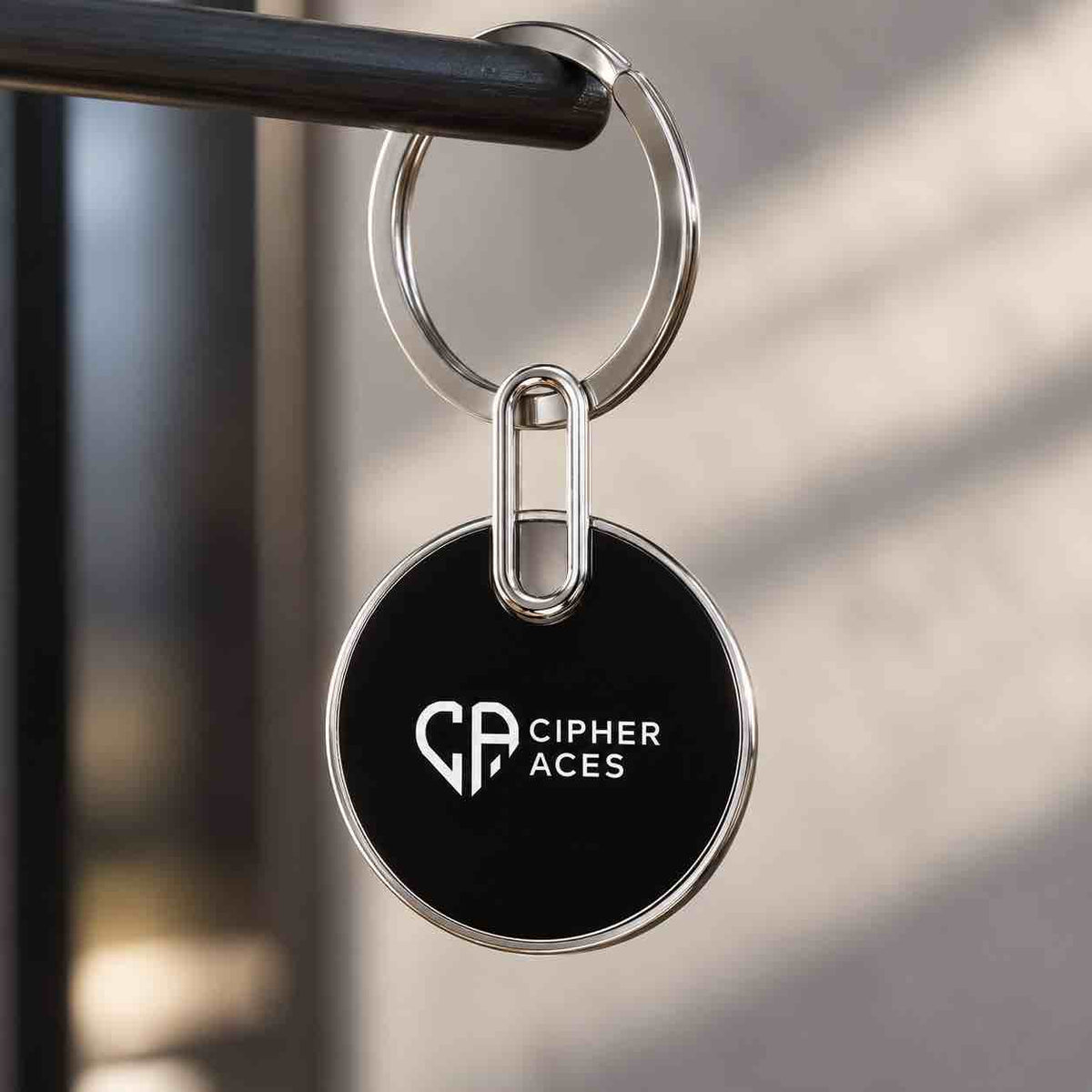 A round black keychain with a silver metal rim and ring, featuring the white Cipher Aces logo, hanging from a dark metal bar against a softly blurred background.