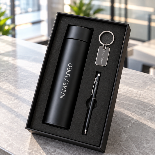 A black gift set in a black box containing a matte black insulated bottle, a black pen, and a silver keychain with a tag, photographed on a gray stone surface with a blurred office background.