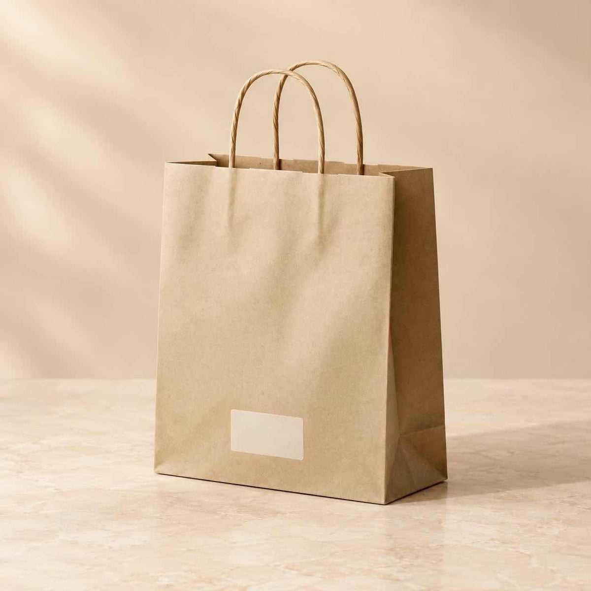 A plain kraft paper shopping bag with twisted paper handles and a blank white label on the front, standing on a light stone surface against a warm beige background.