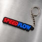 A rectangular keychain with the words 'SPEEDFLOW' in red and blue lettering on a black background, attached to a silver metal split ring and chain, photographed on a brushed metal surface.