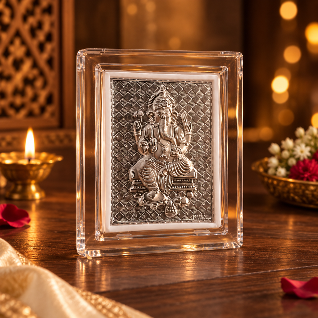A clear acrylic frame containing a detailed silver-toned relief sculpture of Lord Ganesha, displayed on a wooden surface with warm bokeh lights and decorative elements in the background.