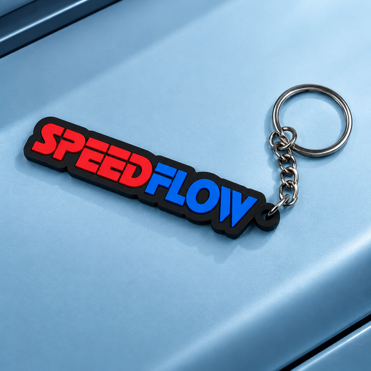 A close-up of a rubber keychain with the word 'SPEEDFLOW' in red and blue letters outlined in black, attached to a silver metal key ring, resting on a light blue metallic surface.