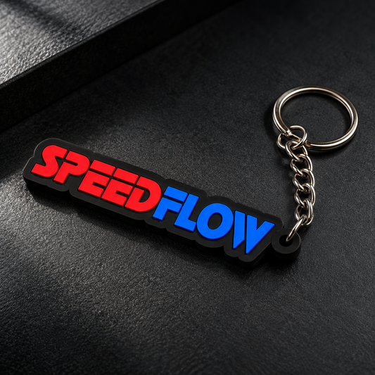 A black rubber keychain with the word 'SPEEDFLOW' in red and blue letters, attached to a silver metal split ring, resting on a dark textured surface.