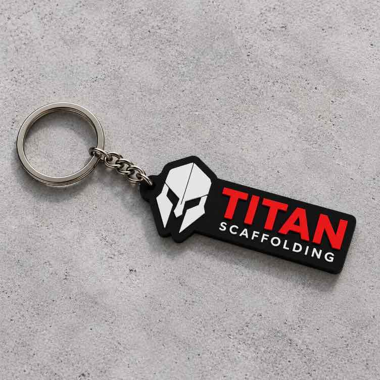 Titan Scaffolding custom keychain with white helmet icon, red TITAN lettering, silver chain and keyring on a textured gray concrete surface.