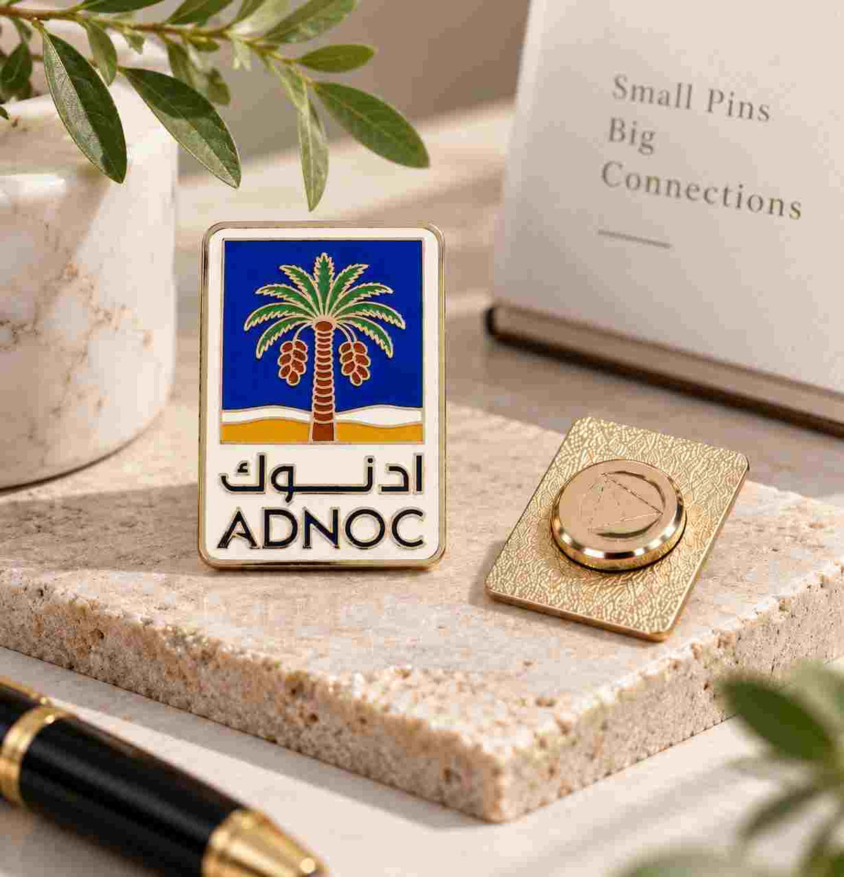 A rectangular enamel pin with a blue and gold palm tree design and the text 'ADNOC' in Arabic and English, displayed on a stone surface next to its gold-toned backing. The scene includes a notebook, pen, and decorative leaves in a styled tabletop setting.