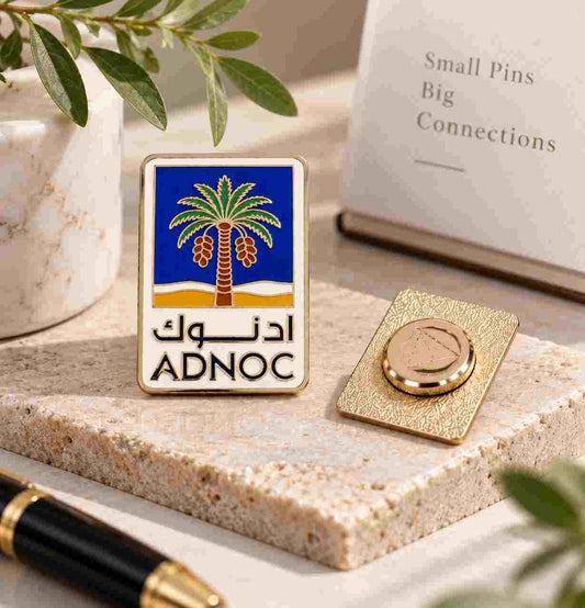 A rectangular enamel pin with a blue and gold palm tree design and the text 'ADNOC' in Arabic and English, displayed on a stone surface next to its gold-toned backing. The scene includes a notebook, pen, and decorative leaves in a styled tabletop setting.