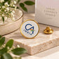 A round enamel pin with a gold border and white face displaying the CA INDIA logo, shown on a stone slab next to its gold clasp and a branded box in a styled setting.