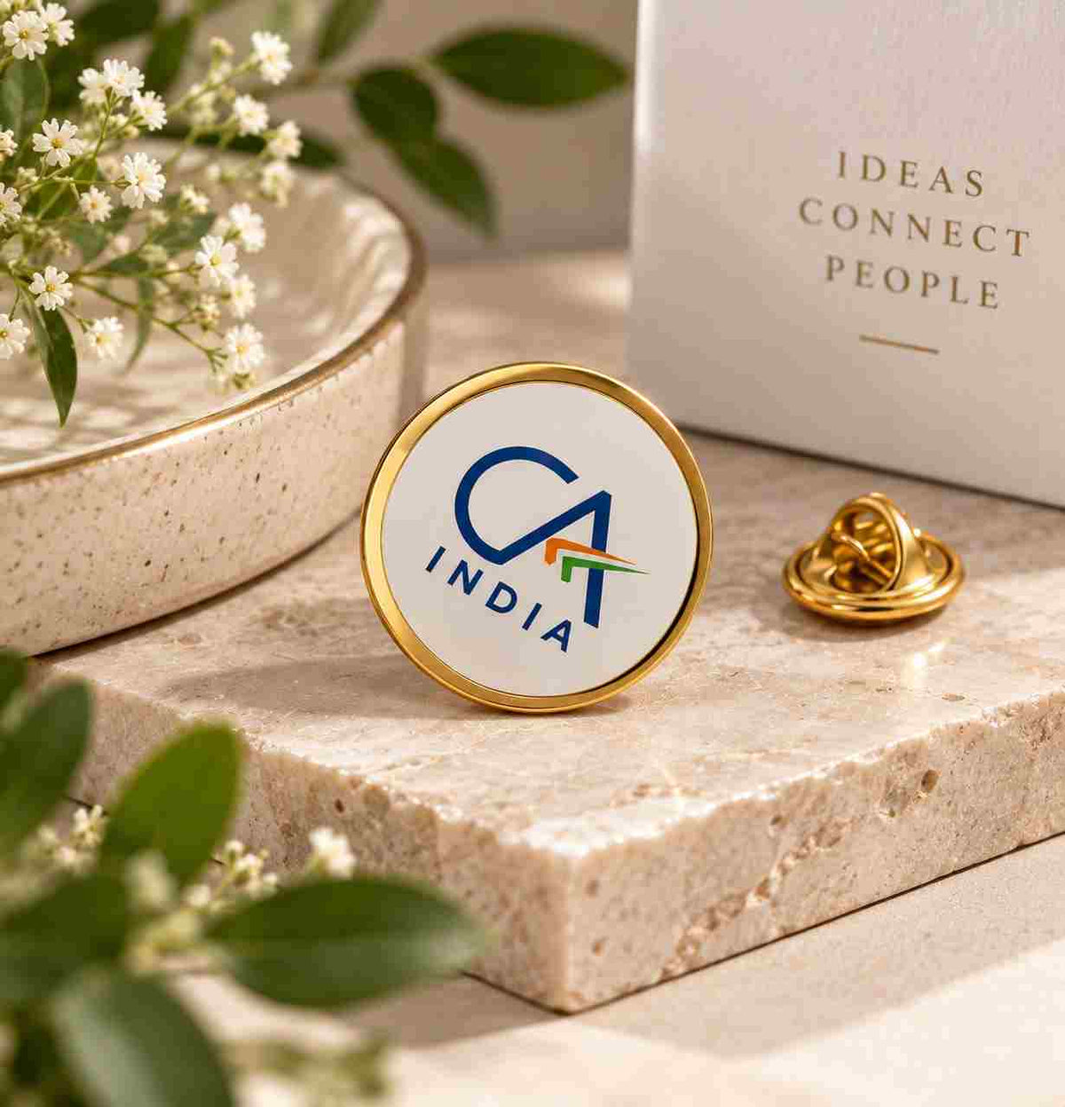 A round enamel pin with a gold border and white face displaying the CA INDIA logo, shown on a stone slab next to its gold clasp and a branded box in a styled setting.