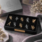A black presentation box containing 12 custom monogram initial studs in gold and silver, styled on a textured mauve fabric with dried flowers and a gray book in the background.