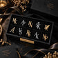 Custom initial brooches designed as elegant alphabet pins, offered in a mix of gold-tone and silver-tone finishes. The pins are displayed in an organized black tray, showcasing the full range of letter options. Styled alongside a luxury gift box and soft floral accents for a premium product presentation. A versatile accessory for personalizing jackets, scarves, or bags.