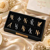 Personalized letter brooch pins arranged in a black velvet box, featuring bold initial designs in gold and silver. Each brooch offers a unique monogram option, making it a thoughtful custom accessory. The elegant styling with dried flowers and a branded gift box reflects a sophisticated, gift-ready presentation. A stylish way to add initials to any outfit or accessory collection.