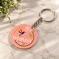 A round acrylic keychain with a coral pink face featuring a colorful hummingbird, a white hibiscus flower, and the text 'WHIMSY Art Studio'. The keychain is displayed on a light marble surface with soft shadows and a blurred plant in the background.