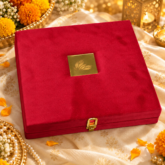 A square red velvet jewelry box with a gold-toned metal clasp and a small gold leaf emblem on the lid, displayed on a cream patterned fabric with decorative flowers and lanterns.