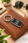 Personalized black metal keychain with silver trim and custom “YOUR LOGO” engraving, styled on a luxury leather notebook.