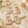 Four customizable gold metal keychains with oval and shield-shaped tags are arranged on a white marble slab with decorative white flowers and a gift box in the background.