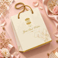A cream-colored paper gift bag with gold rope handles and gold foil accents, styled on a pink satin background with dried flowers and petals.