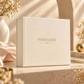 A cream-colored rectangular gift box with 'YOUR LOGO' printed in gold on the front, styled on a beige stone surface with soft sunlight, dried flowers, and a white vase in the background.