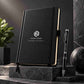 Luxury black personalized notebook and matching pen displayed upright on a dark grey stone pedestal against a charcoal studio background, with dramatic soft lighting and elegant corporate styling.
