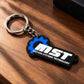 A black and blue rubber keychain with a white 'MST MOTION SYSTEMS TECHNOLOGY' logo, attached to a silver metal split ring, resting on a wooden surface.