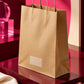 Large brown kraft paper carry bag showcased from a right-side angle on a burgundy pedestal with a hot pink backdrop