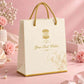 Luxury ivory paper gift bag with twisted gold handles.
Custom logo placeholder printed in gold foil on the front panel.
Elegant script text reading "Your Best Wishes" below the logo.
Photographed against a soft pink floral backdrop.
Perfect for boutique, wedding, or corporate gifting