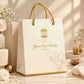 A white paper gift bag with gold twisted rope handles and gold printed logo/text, styled on a marble surface with flowers and soft sunlight in the background.
