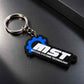 A black and blue rubber keychain featuring the 'MST MOTION SYSTEMS TECHNOLOGY' logo, attached to a silver metal split ring, resting on a dark gray textured surface.