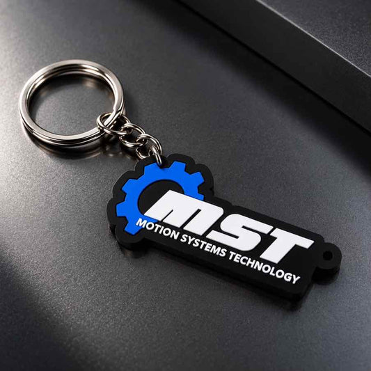 A black and blue rubber keychain featuring the 'MST MOTION SYSTEMS TECHNOLOGY' logo, attached to a silver metal split ring, resting on a dark gray textured surface.