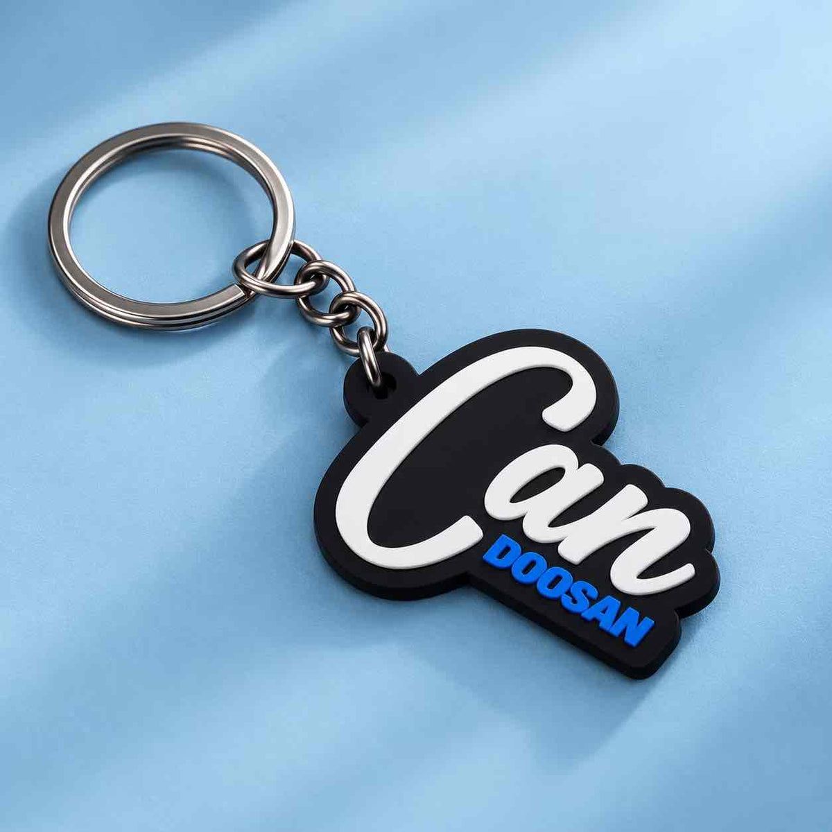 A black and silver keychain with a custom nameplate reading 'Can DOOSAN' in white and blue lettering, shown on a light blue textured background.