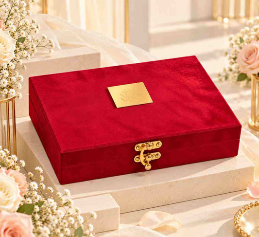 A rectangular red velvet jewelry box with a gold clasp and a small gold plaque on the lid, displayed on a white pedestal with soft floral arrangements in the background.