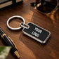 A black rectangular keychain with rounded corners, silver metal border, and a split key ring, placed on a wooden desk. The keychain has 'YOUR LOGO' printed in white.